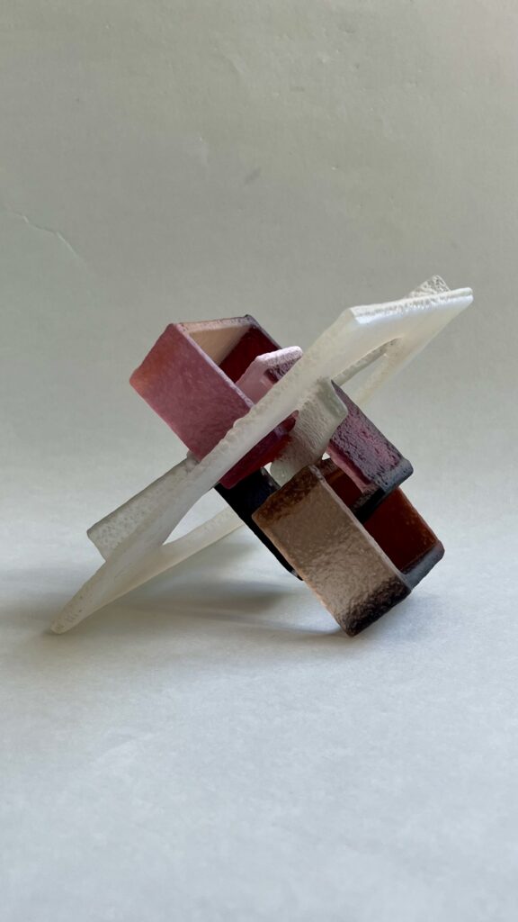 Glass; 3 rectangle shackles  (in pink/brown shades) are melted together with 2 strips on an milky white square with circle cut out of it.